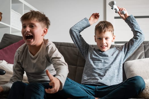 Emotions from winning and losing. School aged boys sit on sofa with joysticks. Children play videogames. Kids having fun in home room at daytime together. Game competition. Best friends lifestyle.