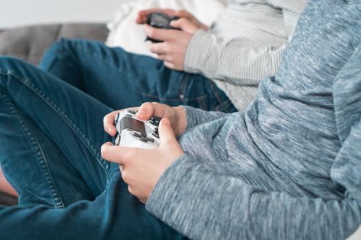 Hands of guys sitting on sofa with game joysticks, closeup side view. Children play videogames. Men in denim and casual longsleeves relaxing at home. Interesting amusement. Best friends lifestyle.