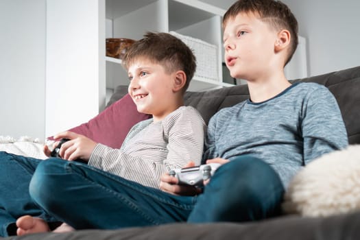 School aged boys sit on sofa with game joysticks, from below view. Children play videogames. Kids having fun in home room at daytime together. Interesting amusement. Best friends lifestyle.