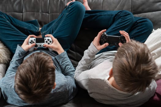 Guys sitting on sofa with game joysticks, from above view of crown and occiput. Children play videogames. Kids in denim and longsleeves relax at home. Interesting amusement. Best friends lifestyle.