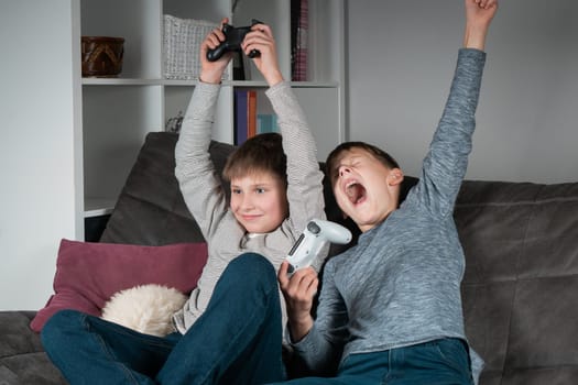 Joyful teenagers sit on sofa with raised arms and game joysticks. Happy boys play videogames. Kids having fun together in home room. Modern electronic toys, winning game. Best friends lifestyle.