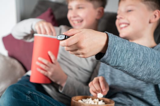 Happy school aged boys sit on sofa, eat popcorn and watch TV. Focus on outstretched hand holding TV remote control. Kids having fun in home room at daytime together. Best friends lifestyle.