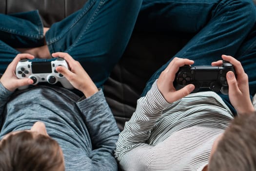 Hands of guys sitting on sofa with game joysticks, from above view. Children play videogames. Kids in denim and casual longsleeves relaxing at home. Interesting amusement. Best friends lifestyle.