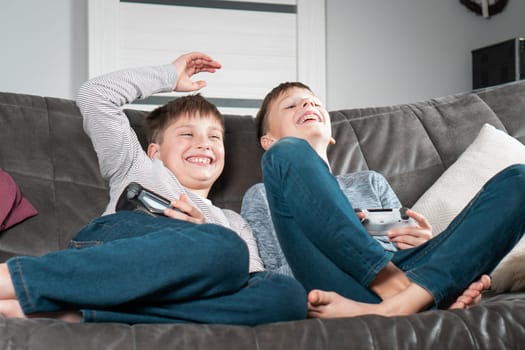 Laughing school aged boys sit on sofa with game joysticks. Children play videogames. Kids having fun in home room at daytime together. Interesting amusement, winning game. Best friends lifestyle.