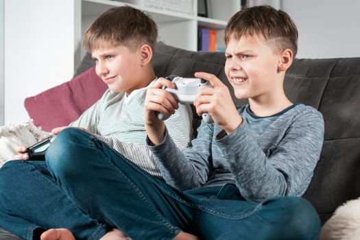 School aged boys in casual clothes sit on sofa with game joysticks. Children play videogames. Kids having fun in home room at daytime together. Interesting amusement. Best friends lifestyle.