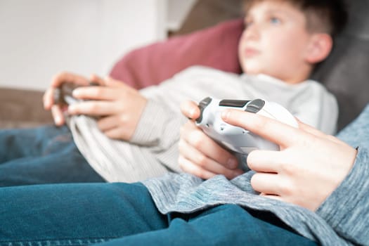 Hands of boy hold modified plastic joystick, selective focus. Children lying on couch play videogames. Hold wireless white game console. Hobby of teenager, lifestyle. Modern technologies and gaming.