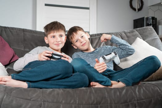 Smiling school aged boys sit on sofa with game joysticks. Children play videogames. Kids having fun in home room at daytime together. Interesting amusement, hobby. Best friends lifestyle.