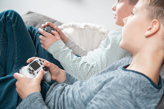 School aged boys sit on sofa with game joysticks, side view from above. Children play videogames. Kids having fun in home room at daytime together. Interesting amusement. Best friends lifestyle.