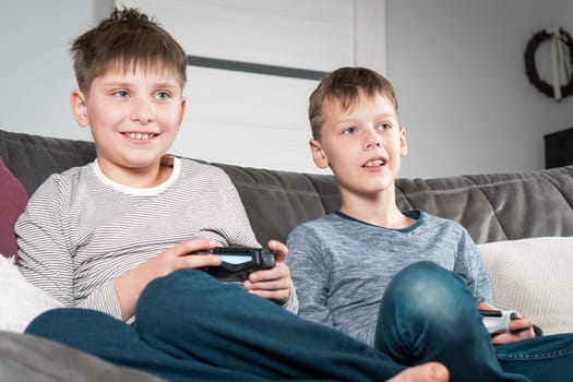 Happy school aged boys sit on sofa with game joysticks. Children play videogames. Kids having fun in home room at daytime together. Interesting amusement, competition. Best friends lifestyle.