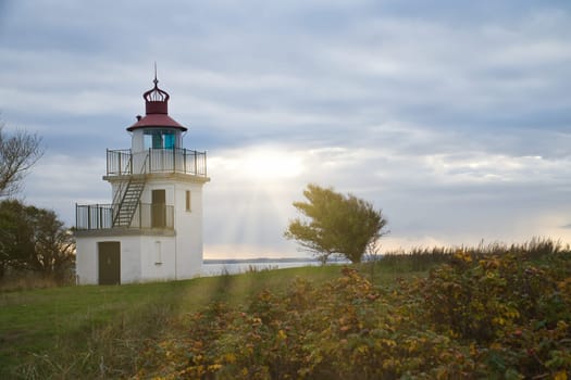 Lighthouse, Spodsbjerg Fyr in Huntsted on the coast of Denmark. Sun rays shining through the clouds. Meadow with trees. Landscape photo from sea