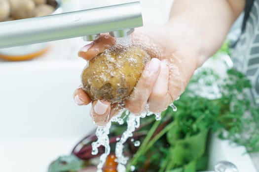 Female hands washing fresh unpeeled potato under tap water jet in kitchen sink closeup. Dirty raw vegetables with green haulm. Cooking, prepare ingredients, food cleaning. Healthy nutrition, diet.