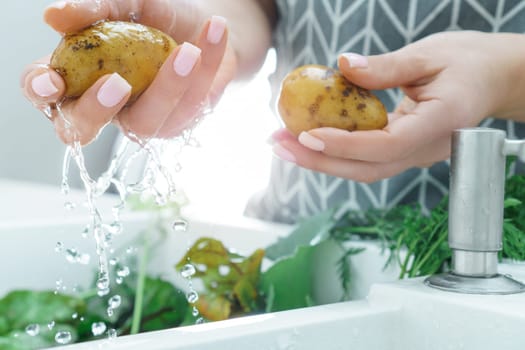 Female manicured hands washing fresh potato under water jet in kitchen sink closeup. Dirty raw vegetables with green haulm. Cooking, prepare ingredients, food cleaning. Healthy nutrition, diet.