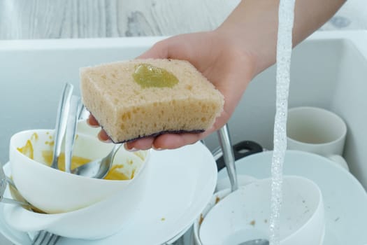 Human hand wash white dinnerware with dish soap on sponge under tap water. Dish washing, tableware after meal, food leftovers. Pile of kitchen utensils in sink closeup. Home cleaning.