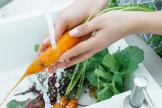 Female hands washing fresh carrot under water jet in kitchen sink closeup. Dirty raw orange carrot with green haulm. Cooking, prepare ingredients, food cleaning. Healthy nutrition, vegetables.