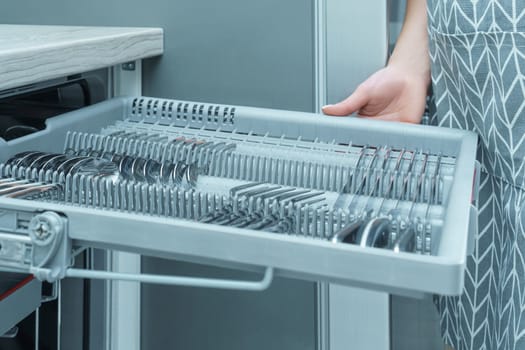 Human hand pull out shelf of dishwasher with clean sparkle cutlery. Dish washing, spoons and forks, household appliance. Modern gray kitchen, silverware cleaning. Convenient kitchen equipment.