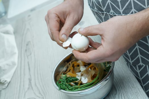 Human hands peeling shell of boiled egg over bowl of vegetable peelings. Person cook on kitchen table, prepare food ingredients. Healthy nutrition, protein product. Separate waste collection.