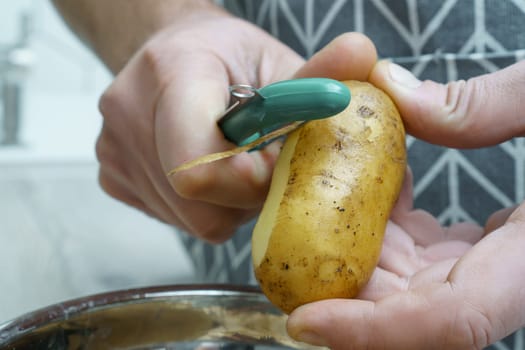 Male hands peeling fresh potato with peeler over bowl of vegetable peelings closeup. Cooking, prepare ingredients, food scrap. Healthy nutrition, vegetables. Separate waste collection, zero waste.