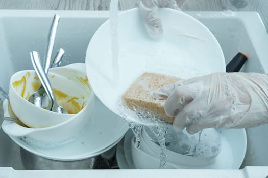 Human hands in protective domestic gloves wash white plate with sponge under tap water. Dish washing, tableware after meal, food leftovers. Pile of kitchen utensils in sink closeup. Home cleaning.