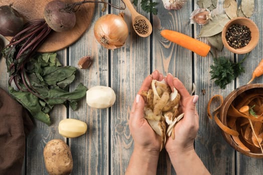 Human hands hold handful of peeled potato peel, flat lay. Mix of raw fresh organic vegetables on wooden table. Healthy food, ingredients for cooking. Zero waste concept. Separate waste collection.