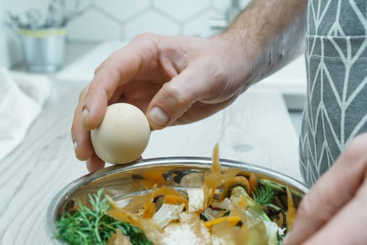 Male hands peeling shell of boiled egg over bowl of vegetable peelings closeup. Cook on kitchen table, prepare ingredients, food scrap. Healthy nutrition, protein product. Separate waste collection.