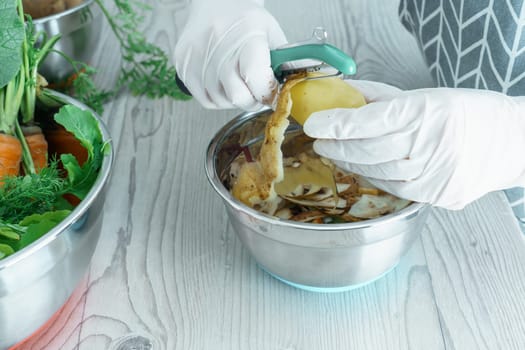 Human hands in white domestic gloves peeling fresh potato with peeler over bowl of vegetable peelings. Cook on kitchen table, prepare ingredients, food scrap. Separate waste collection, zero waste.