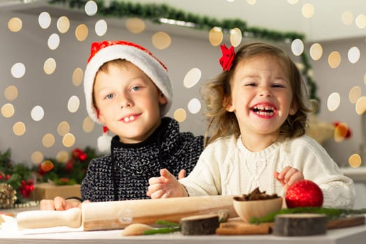 Cute little smiling girl 2-4 with a red bow and boy 7-10 in a Christmas cap, making Christmas gingerbread cookies in the New Year's kitchen.