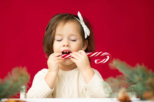 Little girl 2-4 in knitted dress with white bow trying Christmas lollipop with closed eyes on red background. Merry christmas and a happy new year.