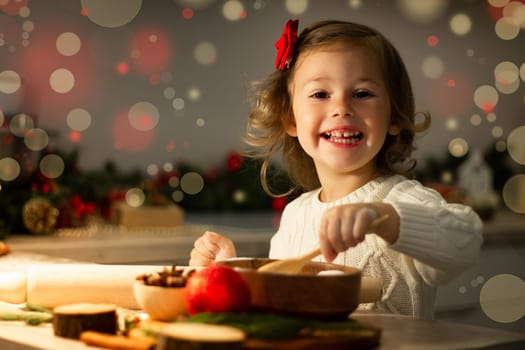 Cute little smiling girl 2-4 with a red bow prepares Christmas gingerbread cookies in the New Year's kitchen.