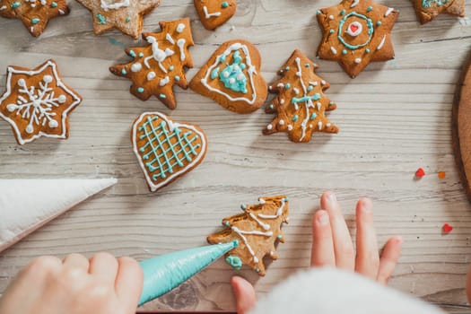 Partially blurred hands of boy decorate gingerbread cookies with blue icing in kitchen. Child prepares cookies on wooden table. Merry Christmas, Happy New Year