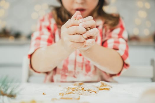 Partially blurred hands of little girl in red checkered shirt prepare dough for gingerbread cookies in white kitchen. Child knead dough on white table strewn with flour