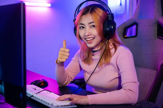 Gamer playing online game wear gaming headphones looking to camera expressing success with game giving thumbs up sign, Smiling woman live stream she play video game at home neon lights living room