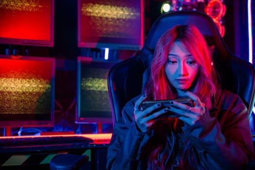 Gamer playing online game application on mobile phone wear gaming headphones, Asian woman live stream she play video game via smartphone at home neon lights living room