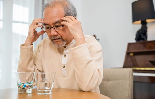 Asian senior old man grey haired sad sitting in chair hand hold head with many medicines and pills for taking, elderly man sick have headache looking pill glass on at home, Elderly healthcare