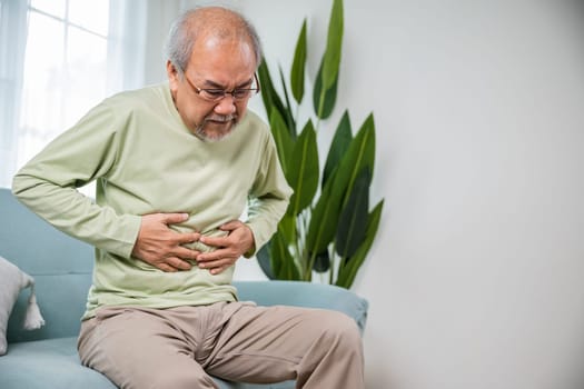 Elderly Asian old man sitting on sofa having stomachache, old age suffering from stomach ache holding his stomach in living room, people health problem, food poisoning