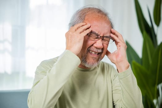 Headache. Close up of elderly holds head with hand suffering from migraine headache, Sad Asian senior man sitting on sofa feeling hurt and lonely at home, Old age health problems, healthcare