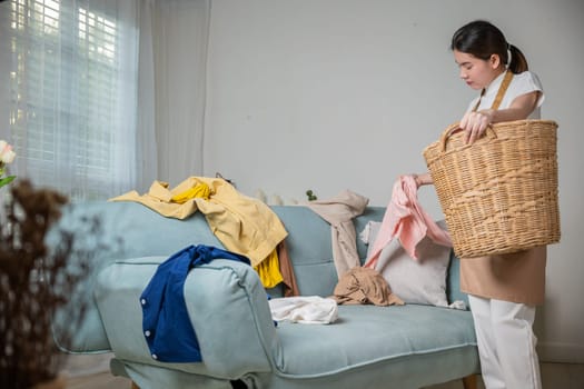 Beautiful young housekeeper maid doing housework holds basket wood of clean messy dirty clothes at home, Housewife woman holding basket with heap of different clothes on sofa, Laundry cleanliness