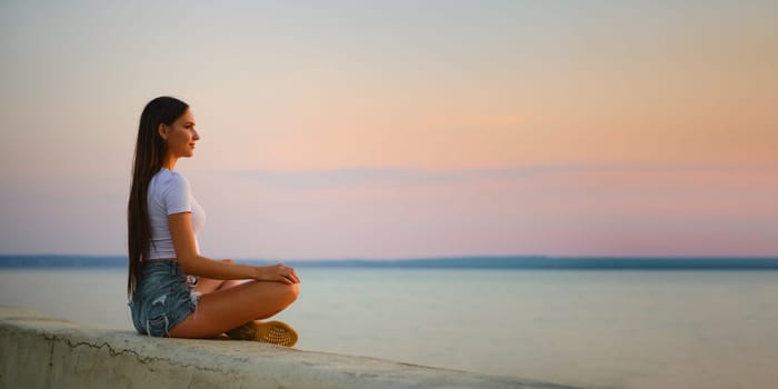 Calm young woman in tank top and denim shorts sits in lotus position on the pier at sunset