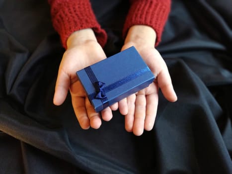 Gift box with a blue bow in children's hands. Black wave-lapped fabric in the background. Gift for New Year, March 8, Birthday, Christmas, Mother's Day or Valentine's Day.