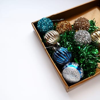 trendy Christmas decorations, colorful balls and tinsel in the lid of a gift box. Shiny beige or brown box for gifts or storage. New Year's Christmas card. Copy space. White background.