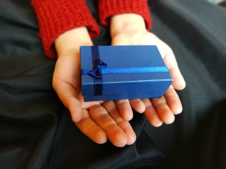 Gift box with a blue bow in children's hands. Black wave-lapped fabric in the background. Gift for New Year, March 8, Birthday, Christmas, Mother's Day or Valentine's Day.