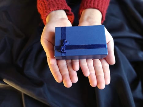 Gift box with a blue bow in children's hands. Black wave-lapped fabric in the background. Gift for New Year, March 8, Birthday, Christmas, Mother's Day or Valentine's Day.