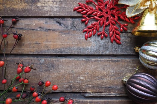 New Year's or Christmas still life. Golden bell. Red fruits of wild rose or rose bush on a wooden background. Yellow and brown striped Christmas balls. Red carved snowflake with sparkles.