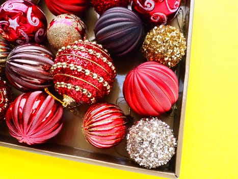 Christmas decorations in a box on a yellow background. Beautiful red and gold balls with sparkles and stripes. Preparing for the New Year Christmas holidays. Beautiful decorations. Copy space