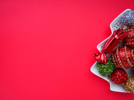 Multicolored Christmas balls and candies on a white plate in the shape of a Christmas tree. Half of the tree is on the right. Copy space Red background. Beautiful striped New Year decor and sparkles.