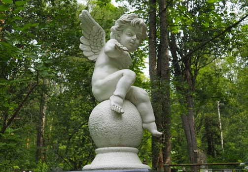 Angel in the cemetery. Monument at the grave of a child. Sculpture in the form of an angel made of marble or plaster against the background of trees in the park. Symbol of love, sadness and death.