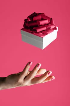 Female hand throwing into air or catching white present box with bow on Color of the Year 2023 Viva Magenta background. Levitating gift. Vertical