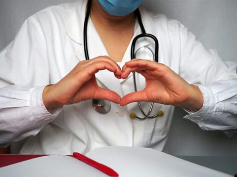 cardiologist doctor in a mask and with a stethoscope shows the heart with his hands. Notebook and pen on the table. Hands gesture. Heart shape made by palms and fingers. Prevention of cardiac disease