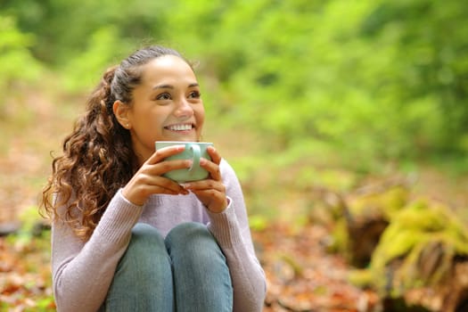 Happy woman sitting in a forest drinking coffee looking away