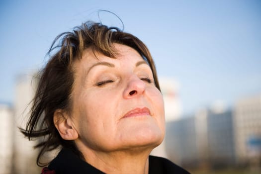 Side head portrait of a mature woman against a fuzzy urban background with closed eyes deeply breathing