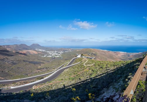 Panoramic view of winding roads in Lanzarote's volcanic landscape, featuring ocean and white villages under a clear blue sky.